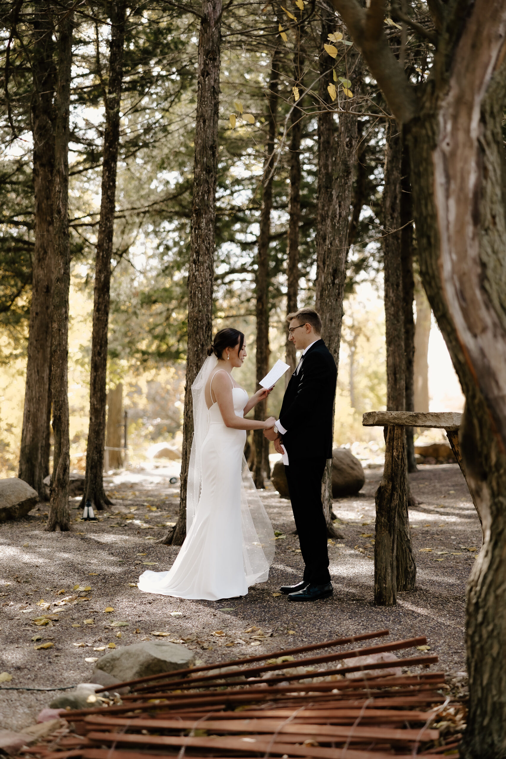 Photograph of a bride and groom reading their vows under tall trees in autumn. This was photographed at Junto Wine in Seward Nebraska by destination wedding photographer Samantha Mudd Photo LLC.